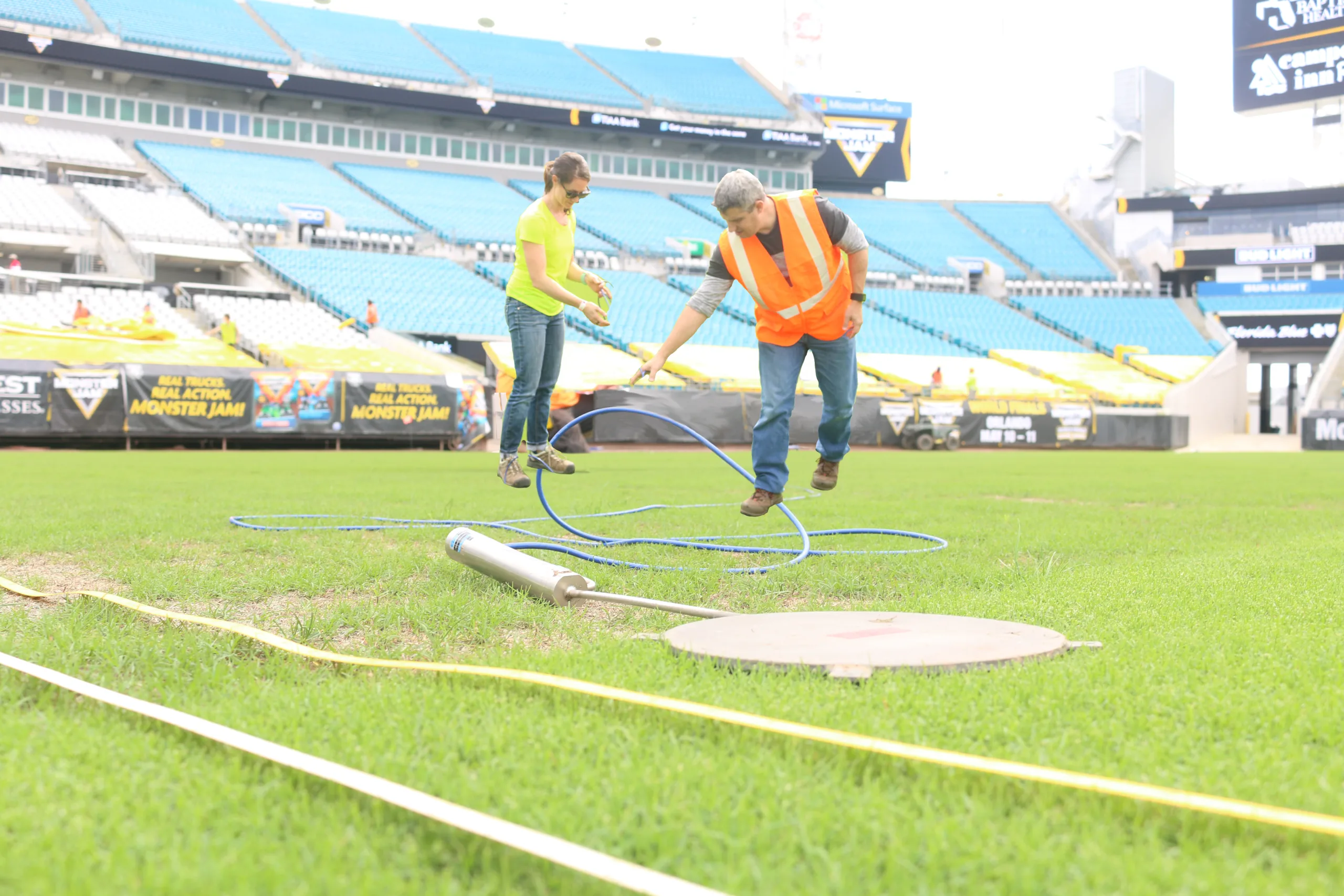 A person in an orange safety vest and a person in a yellow shirt stand on a grass field with blue cables and equipment.