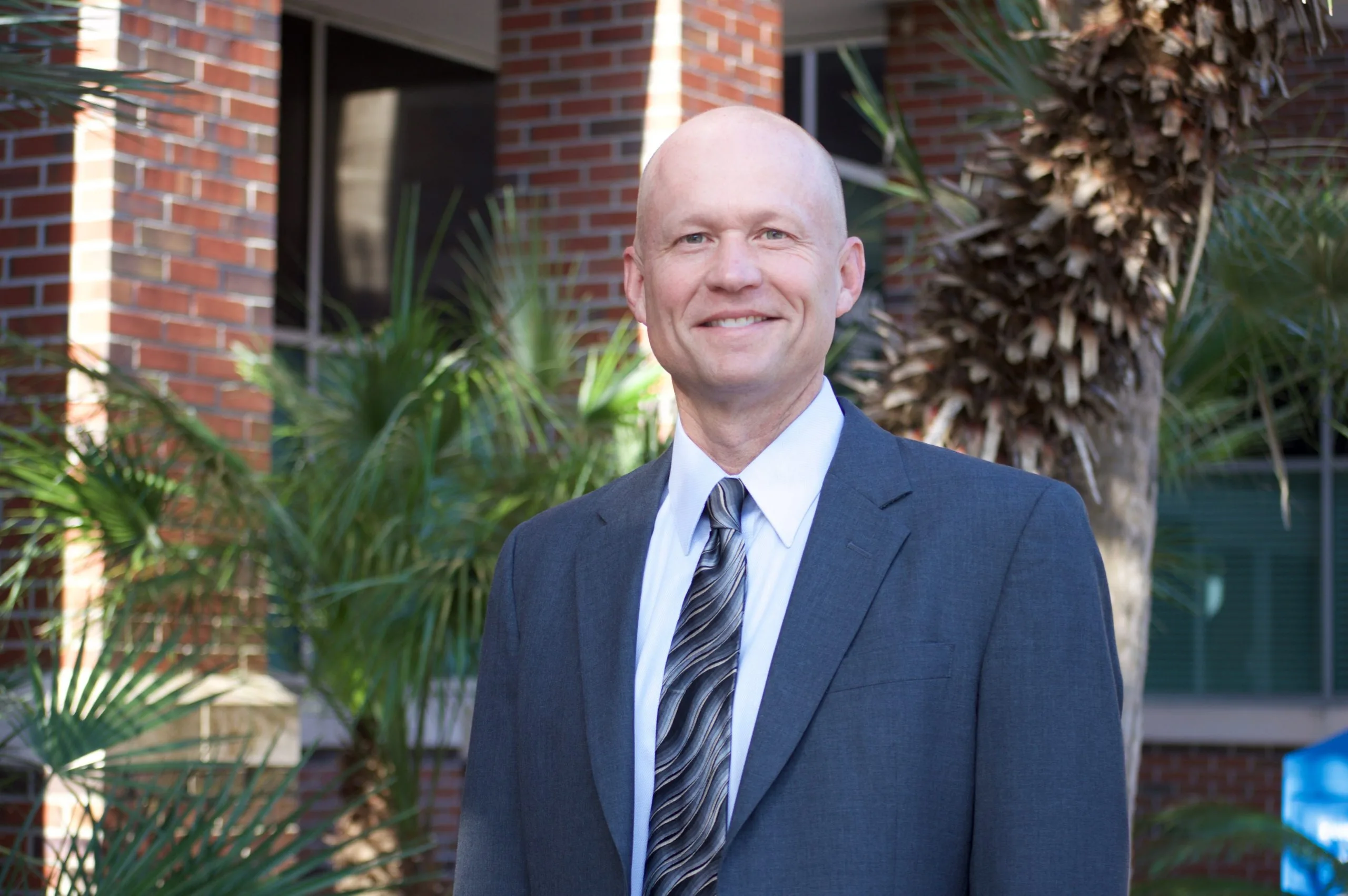 Bald man smiling, wearing a grey suit, light blue shirt, and striped tie, standing before a brick building and palm trees.