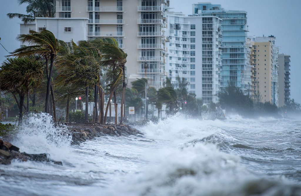 in Florida, Coastal city facing a storm with strong winds, high waves crashing against the shore, and swaying palm trees near tall residential buildings.